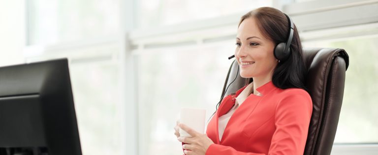 Young attractive woman in red jacket sitting on desk at head of executive in red jacket. Holds video conference in headphones with microphone. Drinking coffee from white cup.