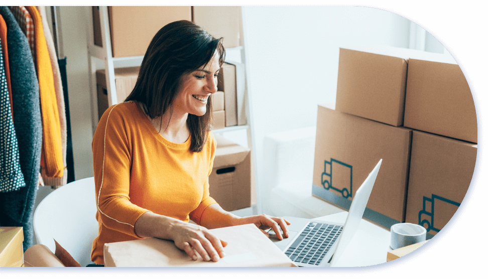 woman sitting at table on laptop with packing boxes in background