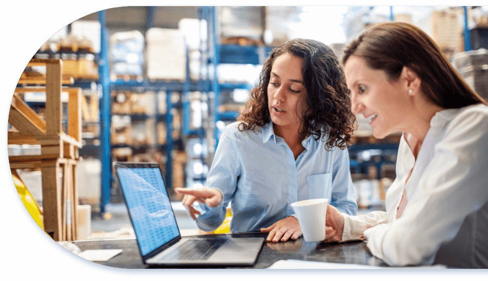 two women sitting at desk talking and using laptop in a warehouse